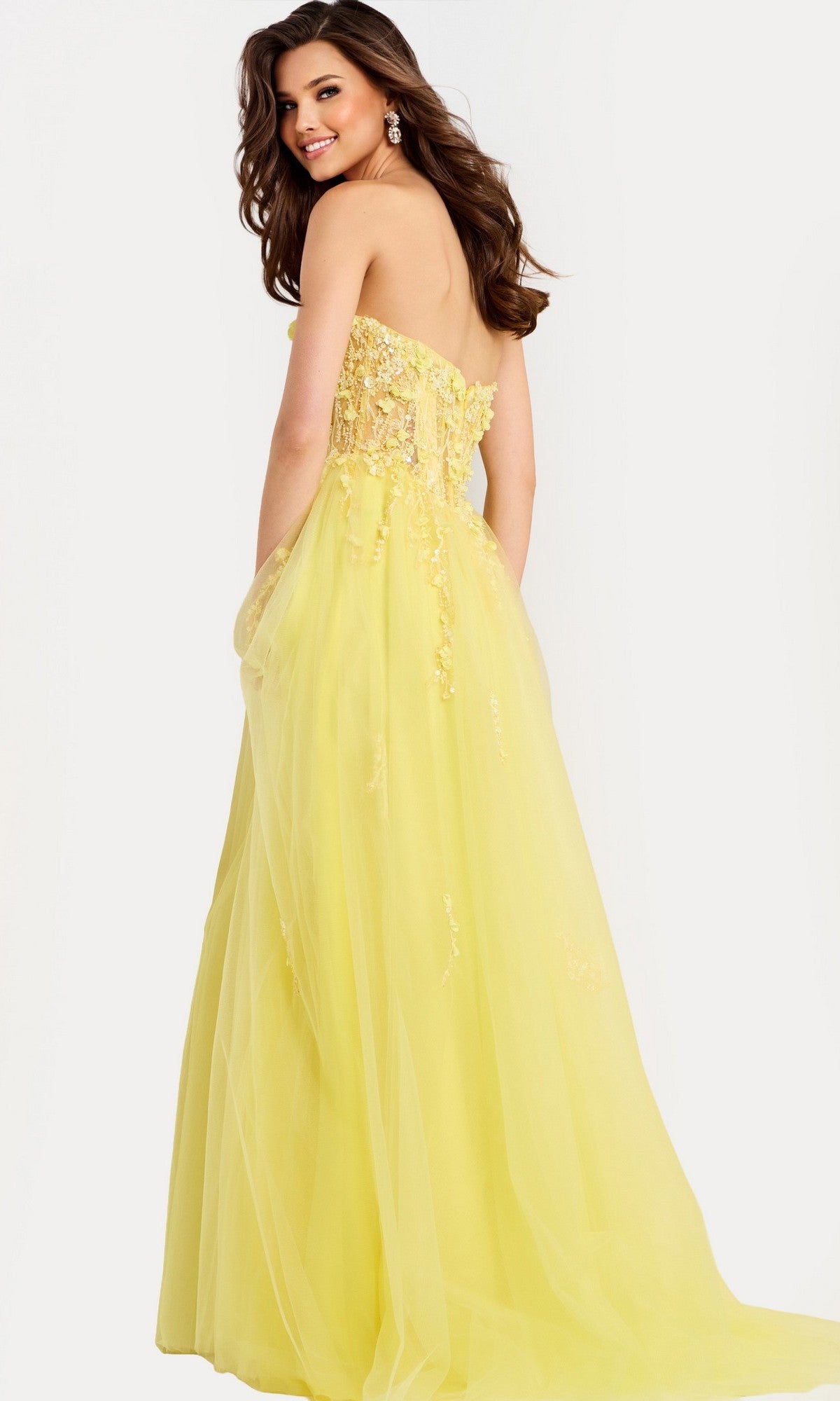 A woman models the Jovani 49704, a long strapless prom dress with a floral-embellished bodice, standing against a plain light background and smiling over her shoulder.